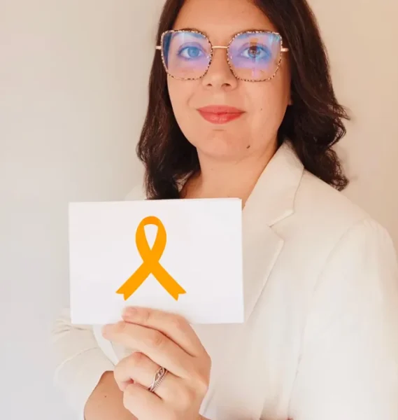 Mujer con gafas sosteniendo una tarjeta blanca con un lazo naranja, simbolizando la concienciación sobre la salud mental, en un entorno neutro.