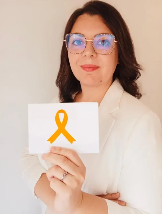 Mujer con gafas sosteniendo una tarjeta blanca con un lazo naranja, simbolizando la concienciación sobre la salud mental, en un entorno neutro.