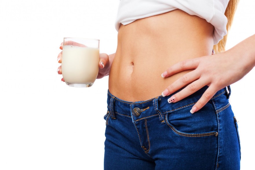Mujer sosteniendo un vaso de leche, mostrando su abdomen, vestida con una camiseta blanca y jeans azules, destacando un estilo de vida saludable.