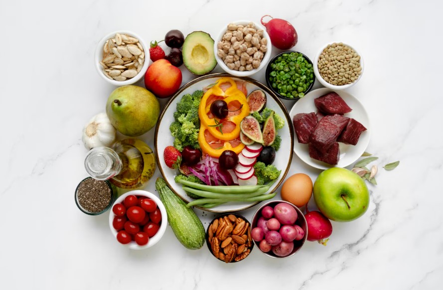 Colorida variedad de alimentos saludables dispuestos en un plato, incluyendo frutas como manzanas, peras, fresas y hortalizas como brócoli, rábanos y pimientos. También se observan legumbres, nueces, semillas y carne magra, todo sobre una superficie de mármol. Ideal para una dieta equilibrada y nutritiva.
